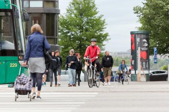 Kävelijöitä ja pyöräilijöitä ylittämässä katua, raitiovaunun kulma näkyy.